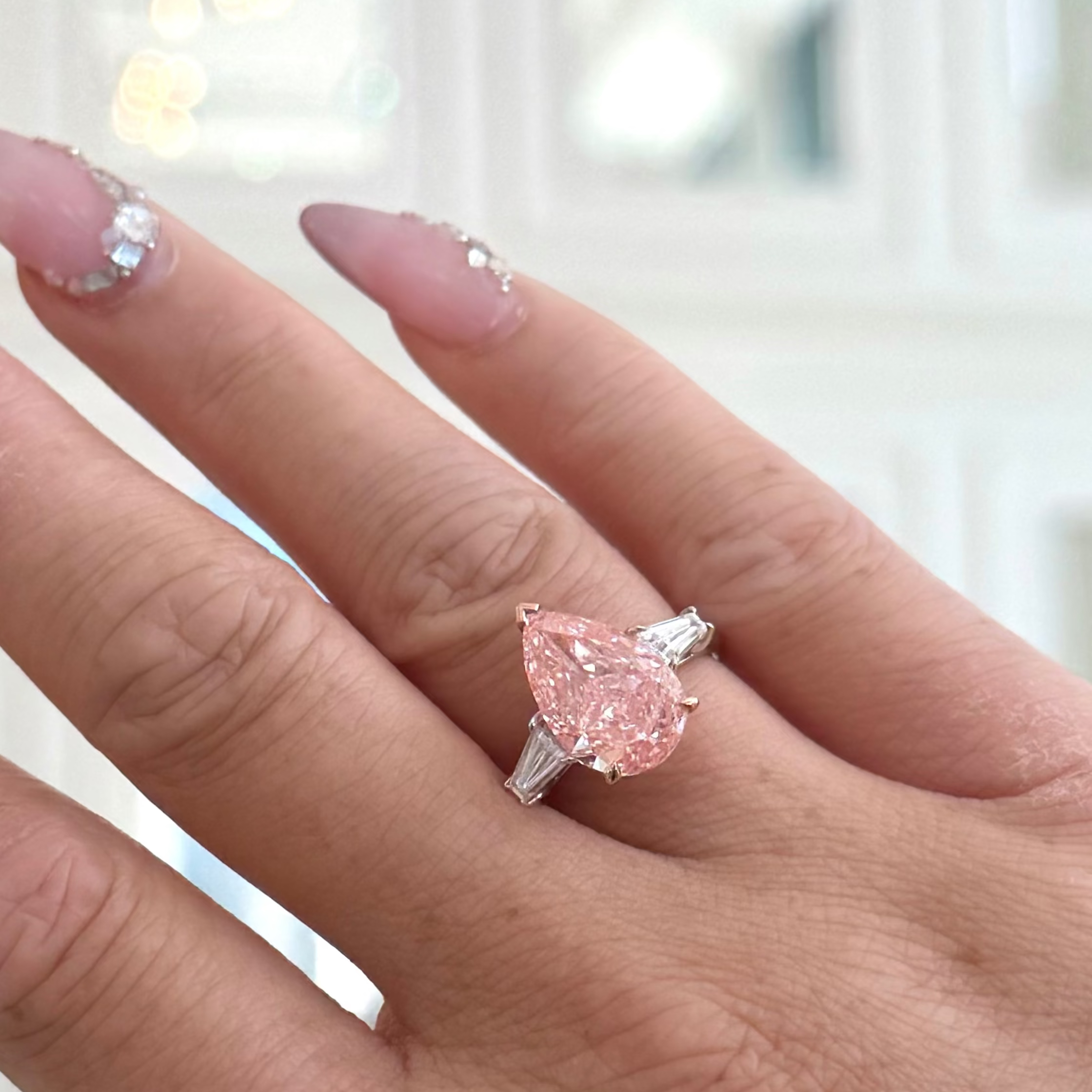 A close-up of a hand wearing a ring with a pear-cut pink gemstone in a silver setting, against a softly blurred indoor background with warm lights.
