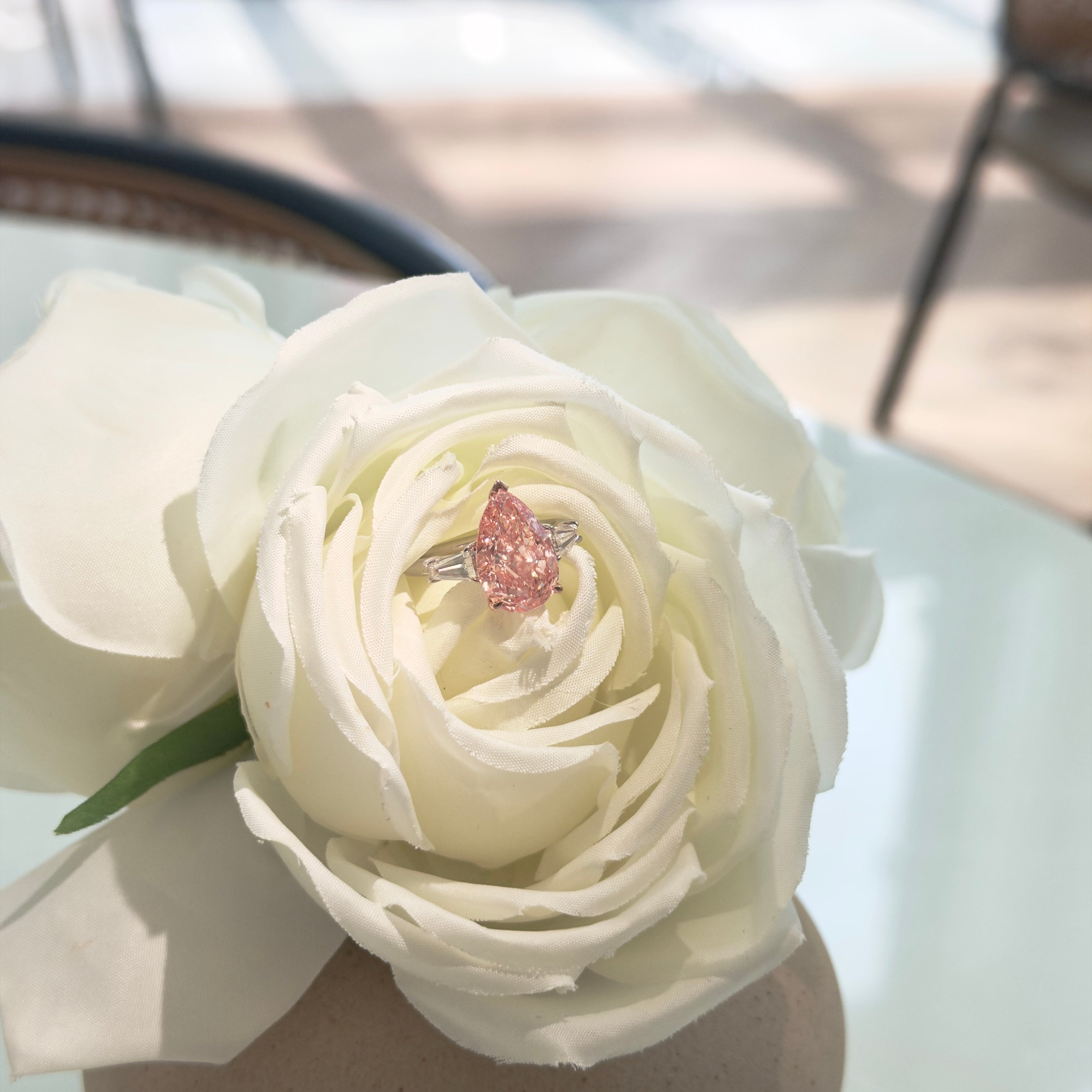 A pink sparkling ring with a pear-shaped center diamond and silver-toned sides, displayed on the tip of a large white rose in a beige vase.