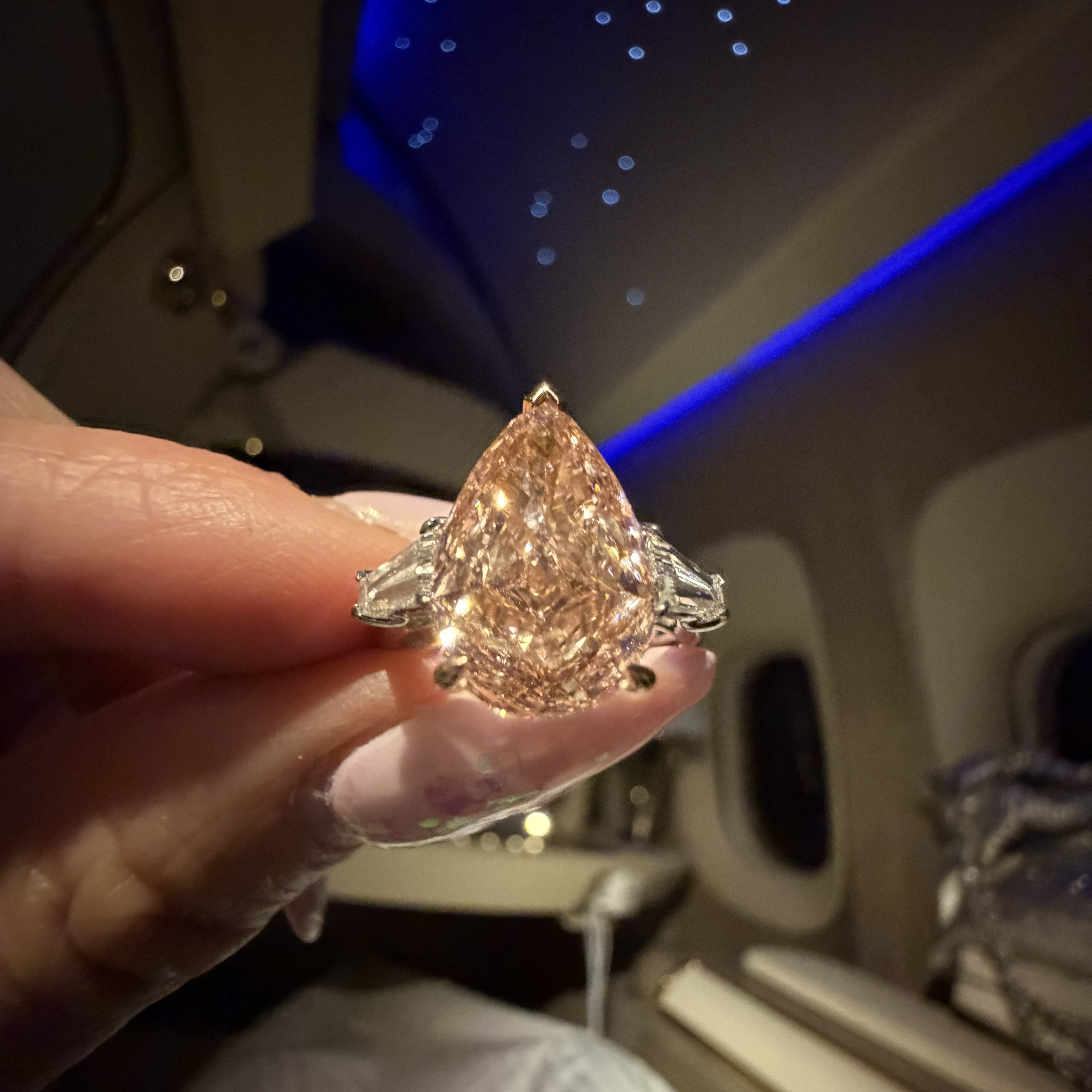 A hand holding a large pear-cut diamond ring with silver-toned side stones, photographed inside an airplane cabin with blue ambient lighting.