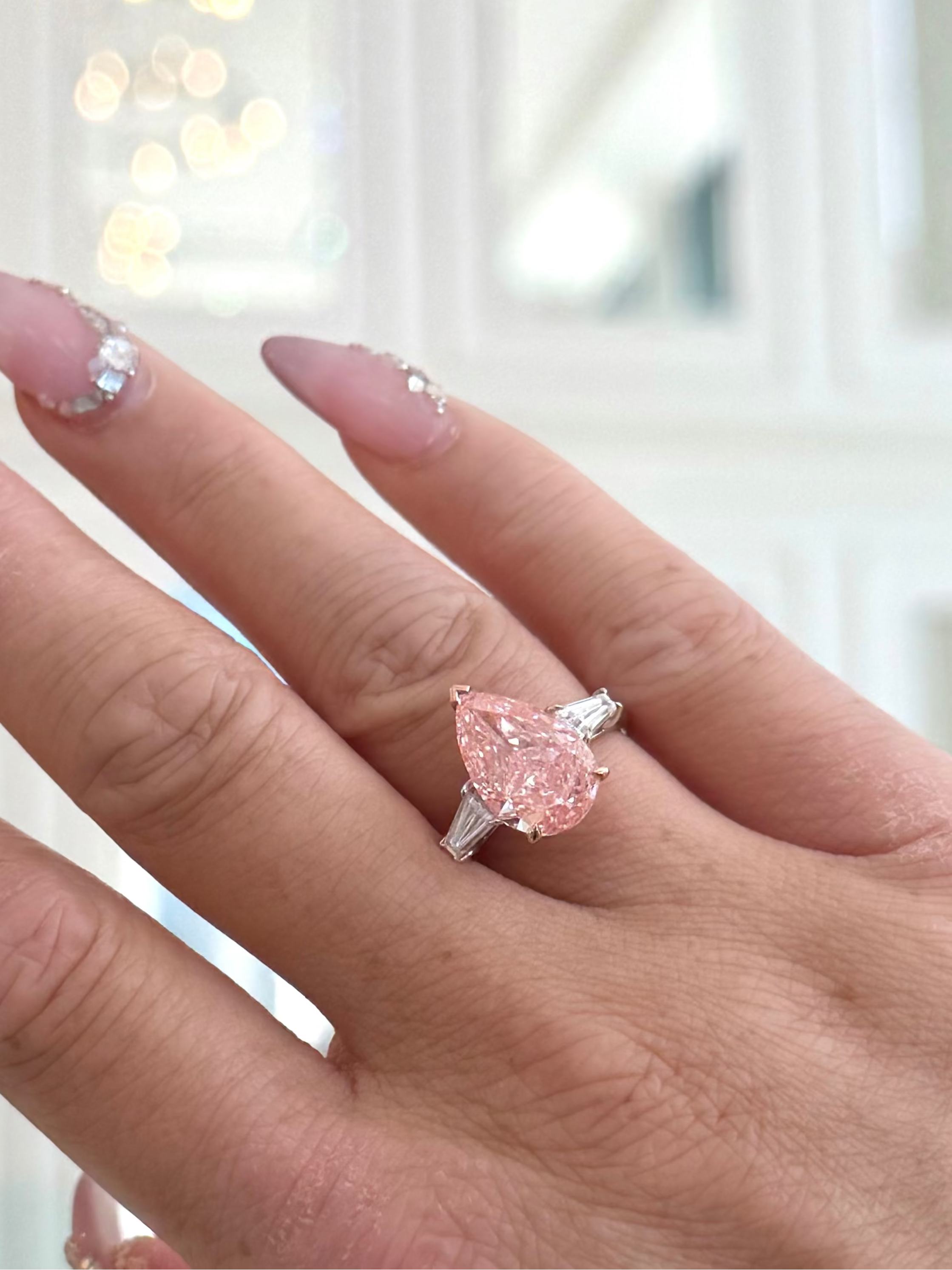A close-up of a hand wearing a ring with a pear-cut pink gemstone in a silver setting, against a softly blurred indoor background with warm lights.