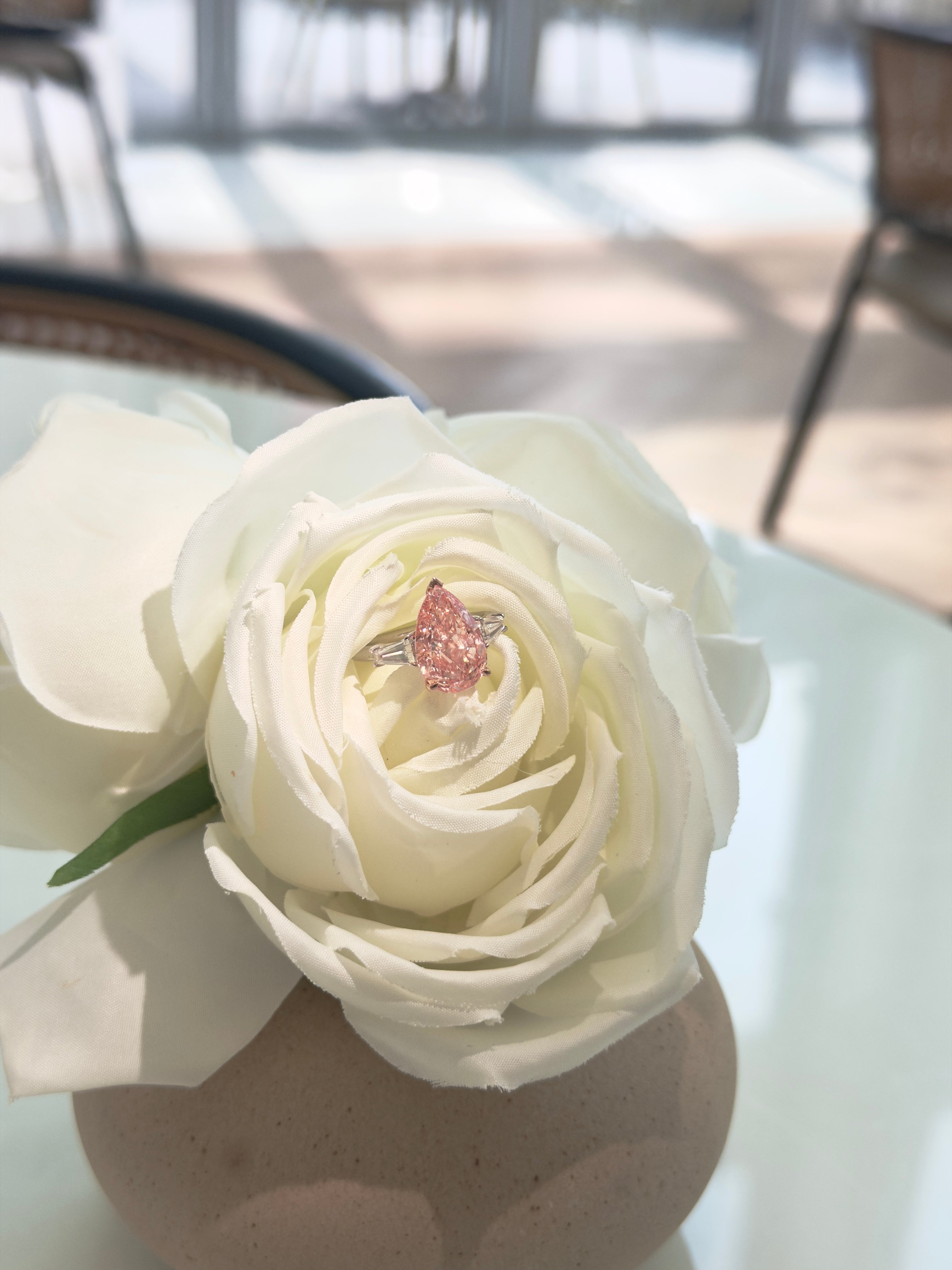 A pink sparkling ring with a pear-shaped center diamond and silver-toned sides, displayed on the tip of a large white rose in a beige vase.