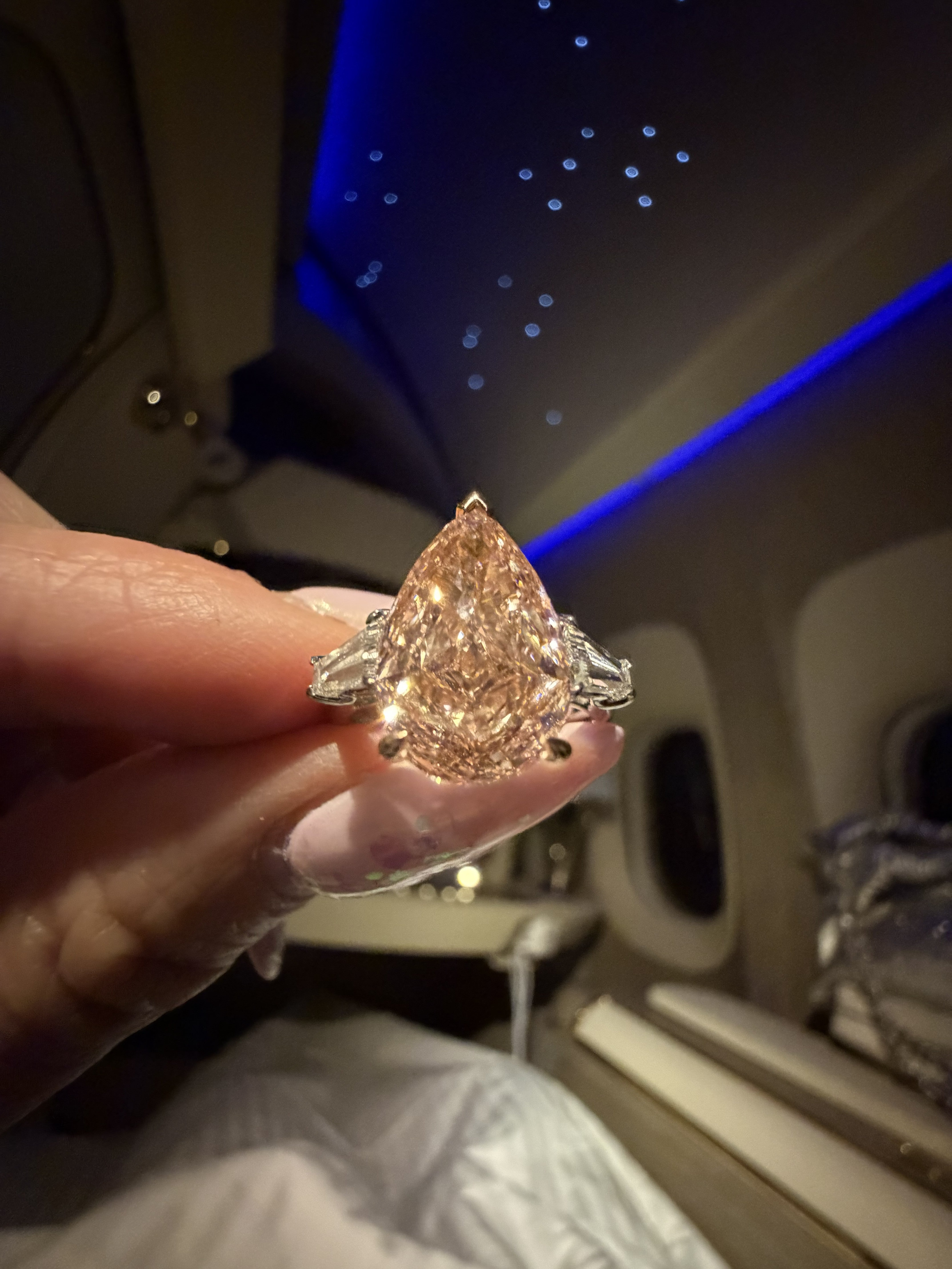 A hand holding a large pear-cut diamond ring with silver-toned side stones, photographed inside an airplane cabin with blue ambient lighting.